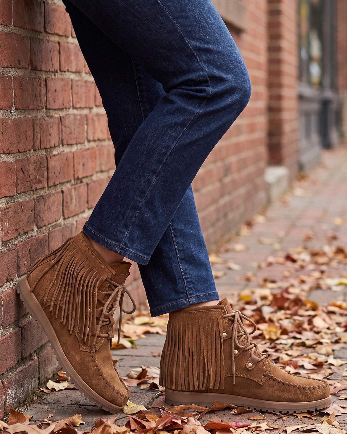 Marlowe fringe moccasin boots worn with jeans, leaning on a brick wall on a leafy sidewalk
